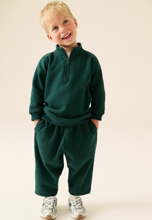 Junger blonder Junge lächelt mit den Händen in den Taschen, trägt einen dunkelgrünen Langarm-Zip-Pullover, passende Hose und weiße Sneakers auf hellem Hintergrund.