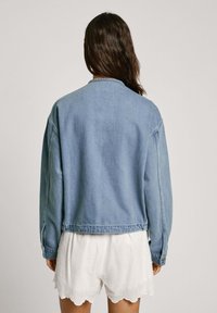 Chaqueta de mezclilla en azul claro, con un ajuste relajado, mangas largas y un cuello de canalé. La vista trasera muestra detalles de costura mínimos y sin cierres.