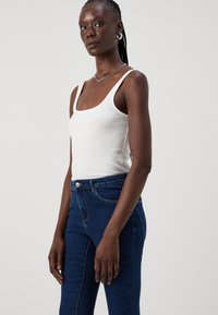 Λευκό ριμπ τοπ με φαρδιές τιράντες, συνδυασμένο με εφαρμοστά σκούρα μπλε τζιν παντελόνια που διαθέτουν μπροστινή τσέπη και μεταλλικό κούμπωμα.