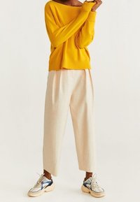 Gelber gerippter Pullover mit einem Off-Shoulder-Design, kombiniert mit hellbeigen weit geschnittenen Hosen und klobigen Sneakers mit blauen Akzenten.