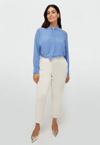 Blusa a maniche lunghe di colore azzurro chiaro con scollatura annodata e dettagli di bottoni sottili, abbinata a pantaloni crema alla caviglia e scarpe argentate con punta.