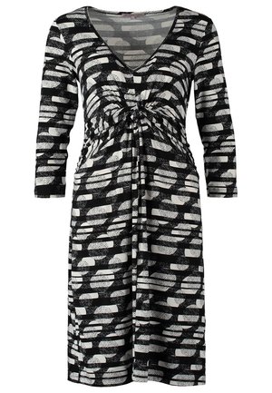 Femme portant une robe noire et blanche à motifs, longueur genou, manches trois-quarts, portant un sac à bandoulière beige et noir et des bottines noires à talons.