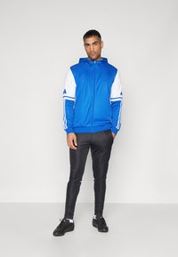Blå hettegenser med glidelås og hvite ermer samt tre-stripe detaljer. Kombinert med svarte tettsittende bukser og svarte treningssko.