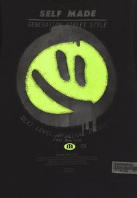 Sudadera negra con un gran gráfico de cara sonriente en neón verde, texto en gris arriba y abajo, y una etiqueta texturizada en la esquina inferior derecha.