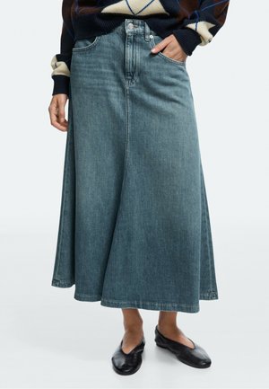 Person wearing a long blue denim skirt, dark sweater with geometric patterns, black flats, one hand in skirt pocket.
