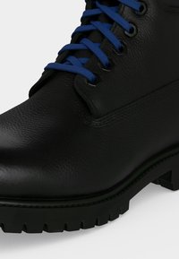 Μαύρο δερμάτινο μποτάκι με υφή, παχύ πέλμα με βαθύ ανάγλυφο και μπλε κορδόνια περασμένα από μεταλλικά ροδέλες, σε κοντινό πλάνο.