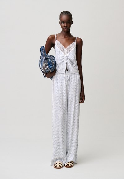 Donna con pantaloni larghi chiari a fiori e top senza maniche abbinato, che tiene una borsa intrecciata blu, in piedi davanti a uno sfondo semplice.