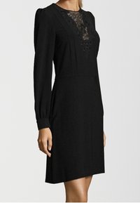 Vestito nero con maniche lunghe, scollatura in pizzo, corpino aderente e gonna a palloncino. Il materiale sembra morbido e texturizzato. I dettagli includono decorazioni in pizzo.