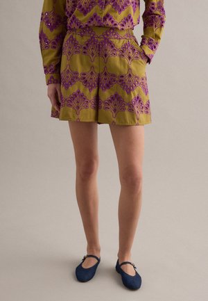 Donna che indossa pantaloncini verde oliva e top abbinato con motivi floreali viola, abbinati a ballerine Mary Jane blu navy. Una mano in tasca.