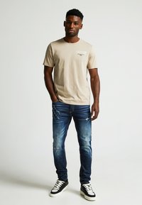 Beige kokvilnas T-krekls ar īsām piedurknēm un grafisku tekstu uz krūšu, kombinēts ar nolietotiem tumši zilajiem džinsiem un melnām sporta apaviem.