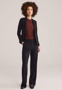 Femeie cu păr creț purtând un sacou negru, pantaloni largi negri, un top de culoare ruginie și pantofi negri împletiți, stând cu mâinile în buzunare.