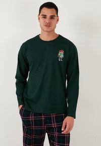Zöld, hosszú ujjú póló, amelyen egy kis, színes mesefigura design található a mellrészen. Az anyag lágynak tűnik, kerek nyakkivágással.