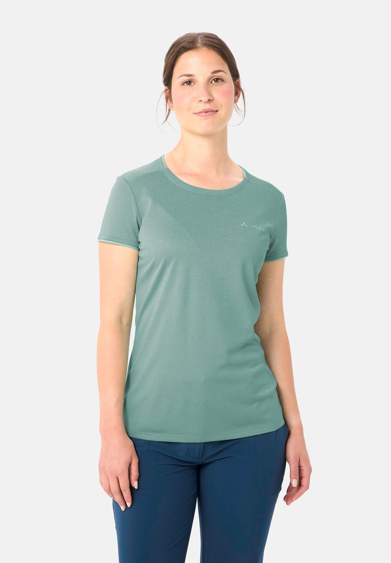 Giovane donna con capelli castani che indossa una maglietta verde chiaro a maniche corte e pantaloni blu scuro, in piedi davanti a uno sfondo bianco semplice.