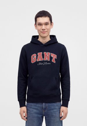 Joven con sudadera con capucha negra con el texto "GANT New Haven" y vaqueros azules, de pie frente a un fondo claro y sencillo.