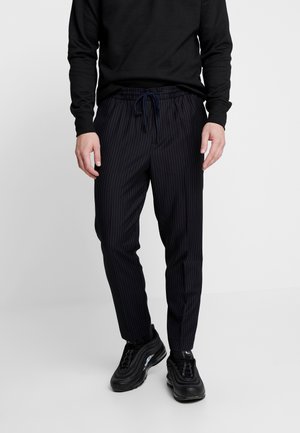 Man draagt een zwart shirt met lange mouwen, zwarte gestreepte broek met trekkoord en zwarte sportschoenen, staand tegen een eenvoudige lichte achtergrond.