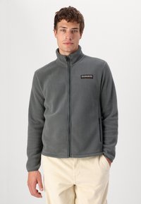 Giacca felpata grigia con zip e colletto alto, dotata di una patch con logo sul lato sinistro superiore, abbinata a pantaloni in corduroy color crema.