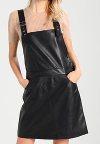 Zwarte leren overall jurk met verstelbare banden, knoopaccenten en zijzakken. Geverfde afwerking en een getailleerde taille.