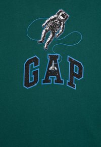 Zelené bavlněné tričko s grafikou astronauta obklopeného modrým pruhem, s nápisem "GAP" v tučných hvězdných písmenech.