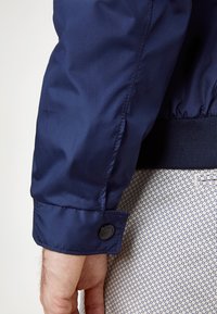 Marineblauwe bomberjack getoond van de mouw naar beneden, met een geribde manchetrand en zwarte drukknopdetail, gedragen over een patroonstof.