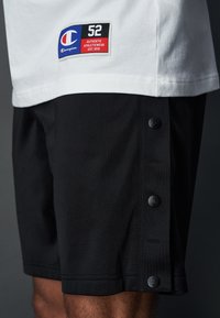 Pantaloni sportivi neri in tessuto testurizzato, con etichetta bianca con logo e numero 52, e sei bottoni a pressione neri sul lato.