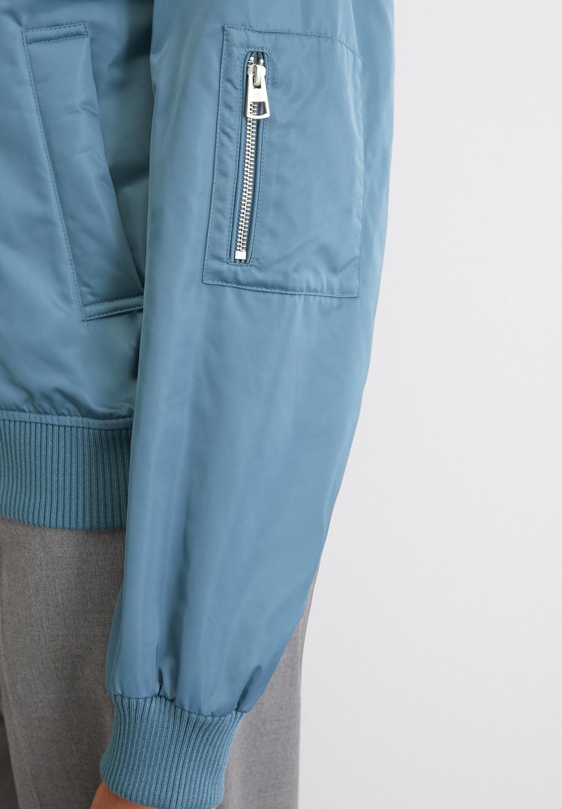 Jasnoniebieska kurtka bomber z prążkowanymi mankietami i dołem, z suwakiem na rękawie oraz gładką, błyszczącą fakturą materiału.