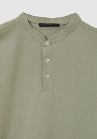 Maglietta henley verde oliva chiaro con scollo tondo, tre bottoni bianchi sul davanti e colletto e polsini rigati. Tessuto morbido e testurizzato.