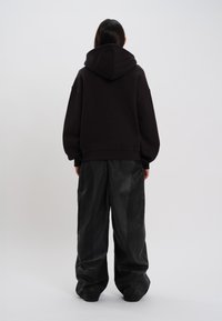 Moletom preto oversized com bolso canguru e um capuz grande, combinado com calças de couro pretas largas, visto de costas.