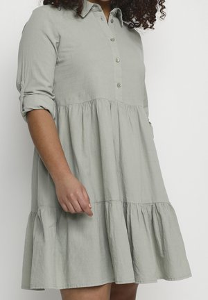 Kvinde iført en lysegrå, knappet, lagdelt kjole med opsmøgede ærmer, der står foran en ensfarvet hvid baggrund.