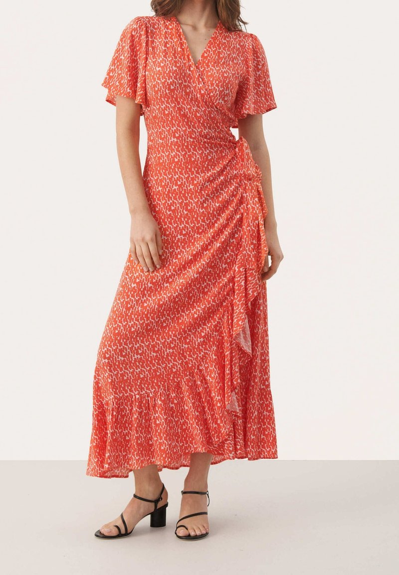 Rotes Wickelkleid mit kurzen flatternden Ärmeln, weißen abstrakten Mustern, geraffter Seite und einem rüschengesäumten Saum, kombiniert mit schwarzen Sandalen mit Absatz.