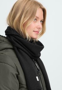 Blond naine, kes kannab oliivrohelist paksu jopet ja paksu musta salli, vaatab paremale väikese naeratusega.
