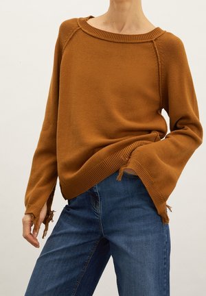 Personne portant un pull en maille marron avec des poignets et un ourlet effilochés, associé à un jean en denim bleu, debout devant un fond uni.