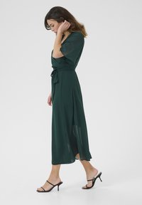 Green, knee-length dress in lightweight fabric with short sleeves and a tied waist. Features a side slit and paired with black heeled sandals.