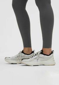 Scarpe da ginnastica bianche con design in rete e dettagli neri, dotate di una suola ammortizzata, abbinate a leggings grigi, presentate su uno sfondo bianco.