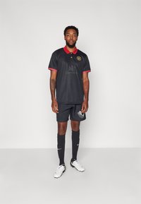 Zwart sporttricot met rode kraag, logo op de borst; bijpassende zwarte shorts; zwarte sokken; witte voetbalschoenen met zwarte accenten; staande pose.
