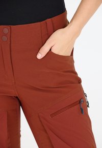 Ruosteenväriset housut, joissa on sileä pinta, etutasku, vetoketjutasku ja kaksipainikkeinen vyötärönauha turvalliseen istuvuuteen.