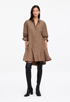 Femme portant une robe marron ample boutonnée avec des manches longues et des bottes noires à talons hautes jusqu'aux cuisses, debout sur un fond blanc uni.