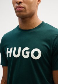 T-shirt verde escuro de algodão, com texto branco em destaque "HUGO" estampado no peito. Design clássico de gola redonda com mangas curtas.