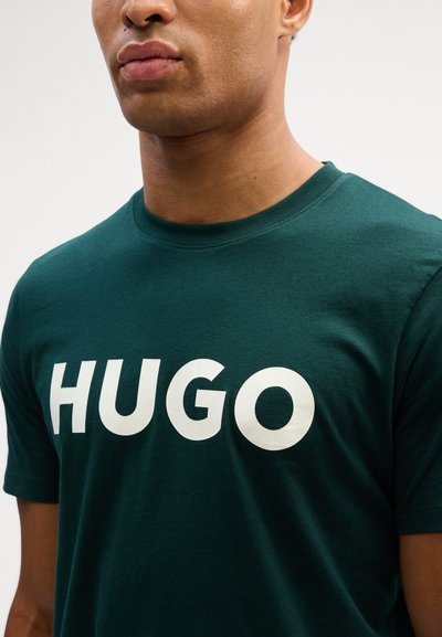 Тъмнозелена тениска, изработена от памук, с ярък бял текст "HUGO" на гърдите. Класически дизайн с кръгло деколте и къси ръкави.