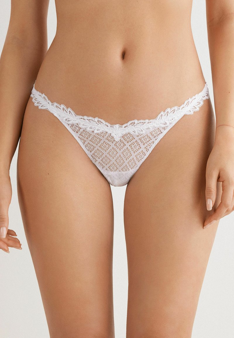 Witte kanten string met een geometrisch patroon en golvende randen. Zacht, lichtgewicht materiaal; heeft een delicate bloemenkantrand langs de tailleband.