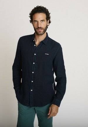 Homme aux cheveux bouclés et à la barbe, portant une chemise bleu foncé boutonnée et un pantalon vert, debout devant un fond clair uni.