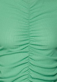 Top verde ajustado com plissado vertical na frente, em um tecido leve e texturizado. O design em V realça sua estrutura.