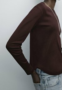Maroon long-sleeve shirt with a rounded neckline and side slits, paired with light blue denim jeans. Soft fabric, form-fitting design.