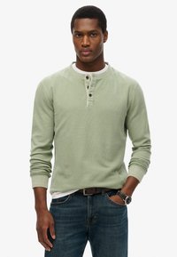 Camicia henley verde a maniche lunghe con finitura testurizzata, placca con tre bottoni e polsini a coste abbinata a jeans scuri.