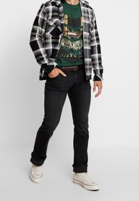 Langärmliges schwarz-weiß kariertes Hemd über einem grünen Grafikshirt, kombiniert mit schwarzen Jeans und weißen Sneakers. Lässiger Look.