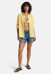 Geel-witte gestreepte button-up shirt, denim shorts en zwarte sandalen. Het shirt heeft opgerolde mouwen en een ontspannen pasvorm.