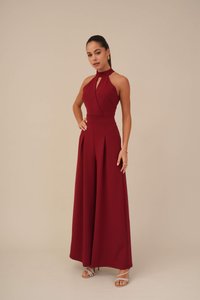 Combinaison dos nu bordeaux avec décolleté en trou de serrure, jambes larges plissées, fabriquée en tissu lisse, associée à des sandales à lanières argentées.