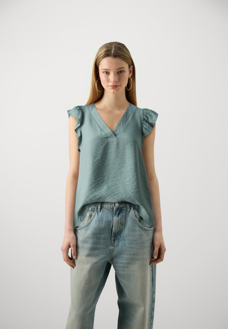 V-neck, short-sleeved blouse in light teal fabric with a textured finish and ruffled sleeves, paired with high-waisted light blue jeans.
