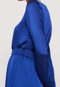 Robe bleue à manches longues avec une finition texturée semblable à du satin. Dotée d'une taille smockée et d'une coupe décontractée.
