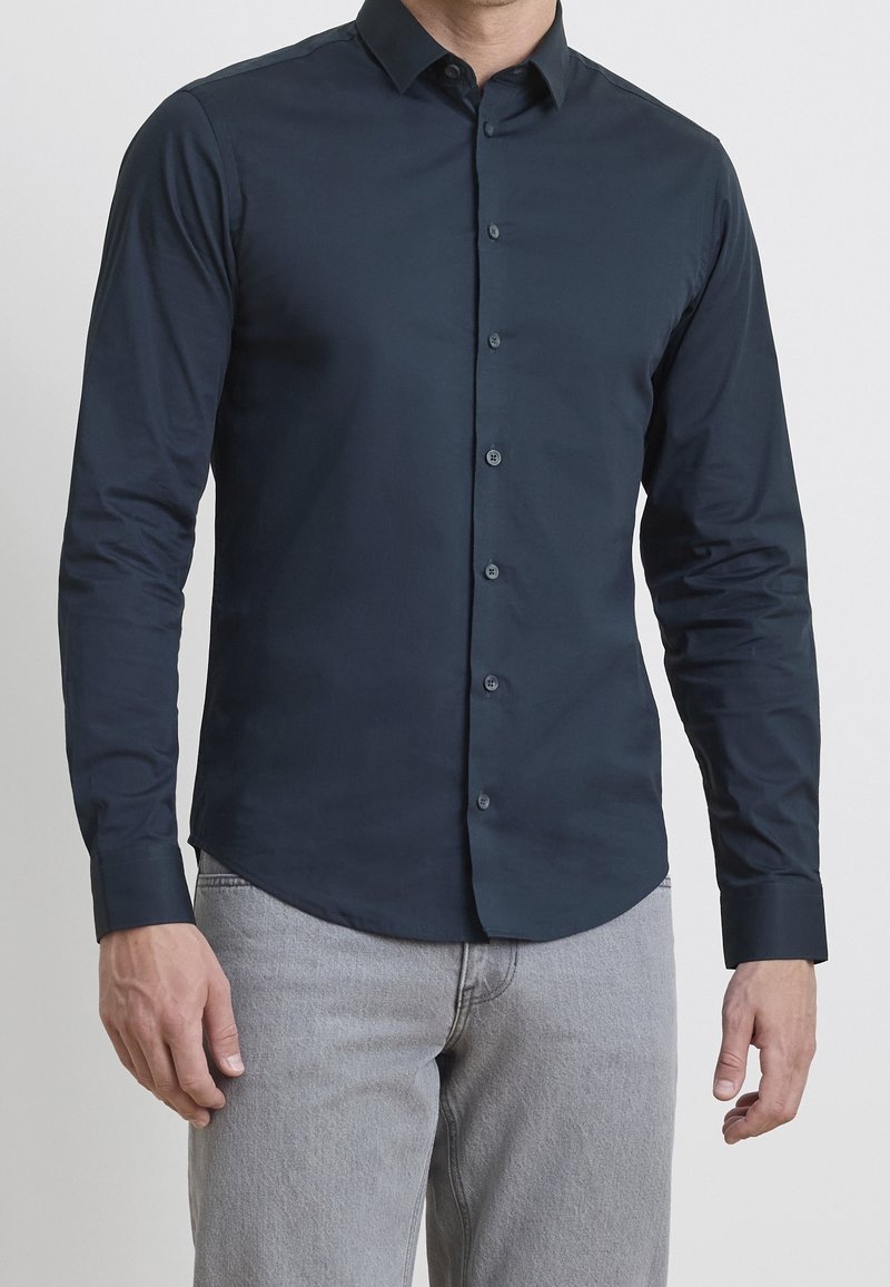 Homme portant une chemise ajustée à manches longues bleu foncé à boutons et un jean gris clair, debout devant un fond uni.