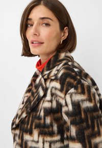 Wollen blend jas met een geometrisch patroon in bruin, crème en zwart. Opvallende details zijn onder andere een gekruist revers en een zachte textuur.